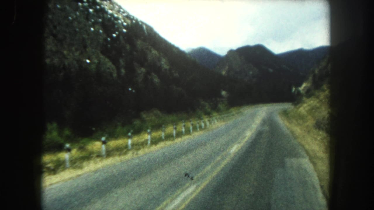 Vehicle POV of a winding Summit County mountain road, with roadside posts and forested ridges visible in soft 8mm color.