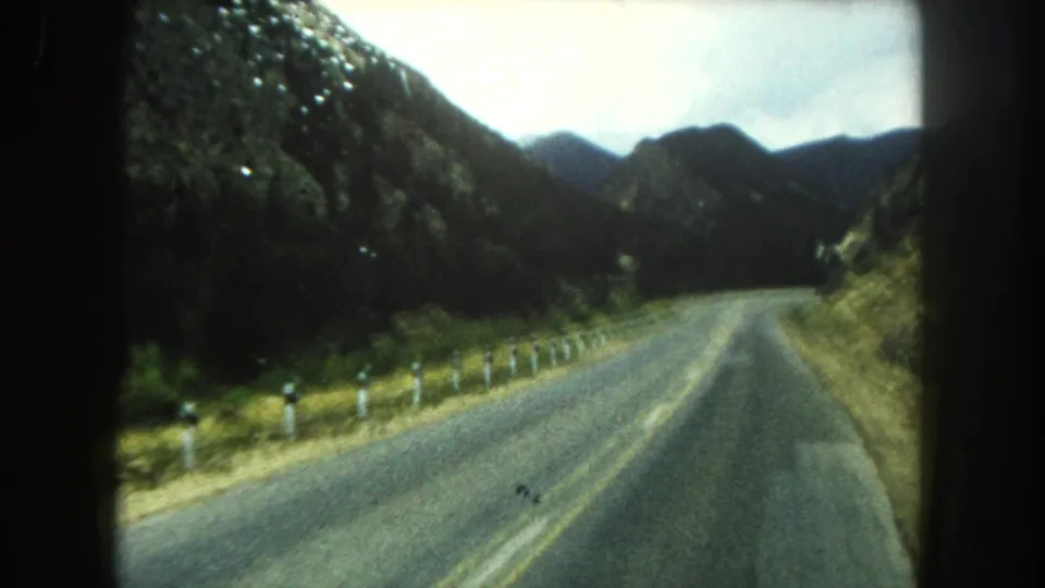 Vehicle POV of a winding Summit County mountain road, with roadside posts and forested ridges visible in soft 8mm color.