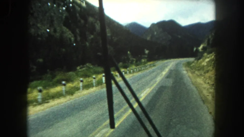1962 home-movie road view through a vehicle windshield, with wiper blades visible over a two-lane road in Colorado mountains.
