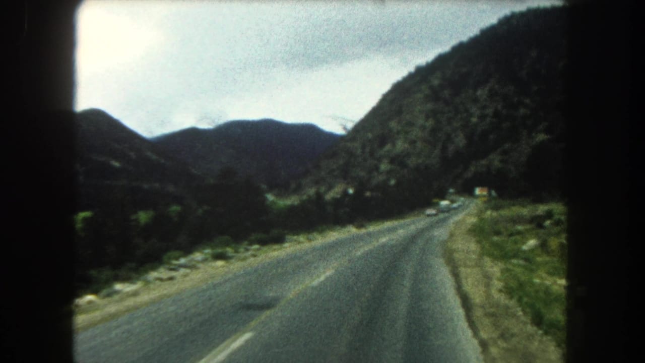 1962 8mm view from inside a car on a Summit County mountain highway, with road, hills, and overcast sky ahead.