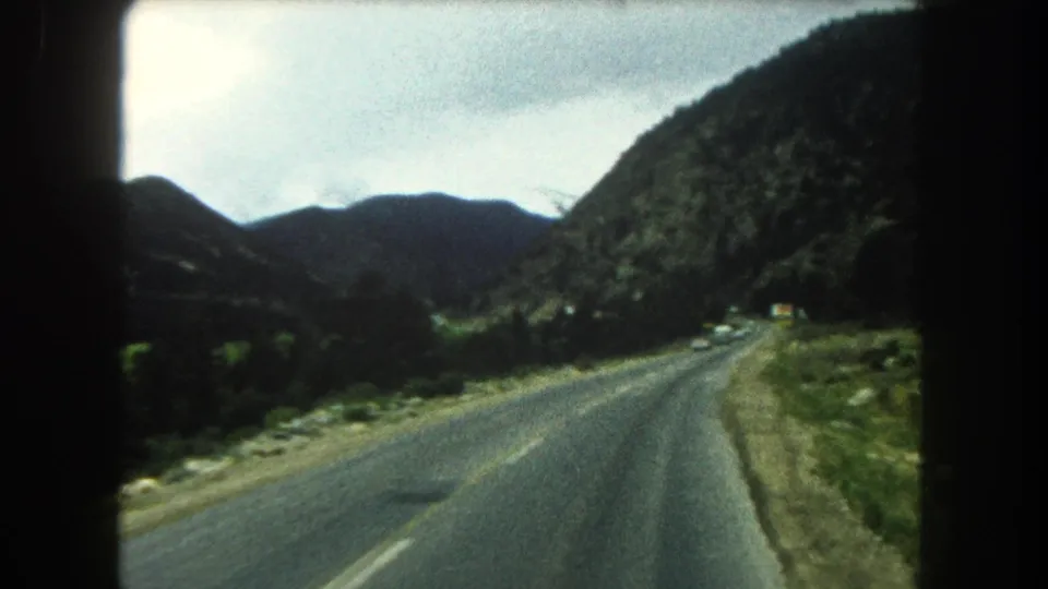 1962 8mm view from inside a car on a Summit County mountain highway, with road, hills, and overcast sky ahead.