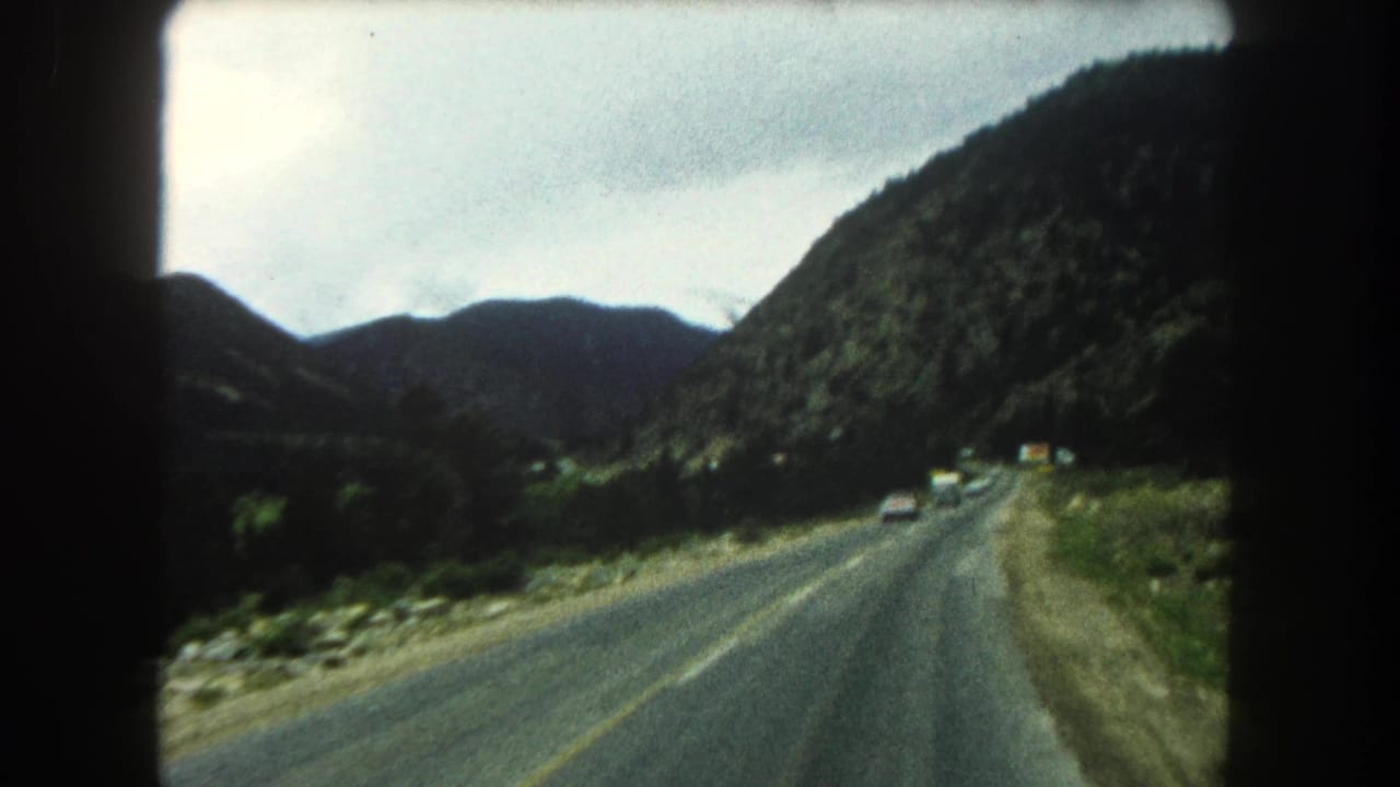 Mountain road with cars ahead, rocky roadside, forested hills, and overcast sky.
