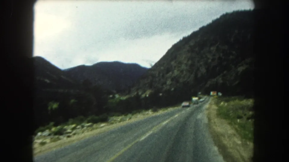 1962 Summit County road-view footage shows cars ahead on a two-lane highway bordered by rocky ground and wooded hills.