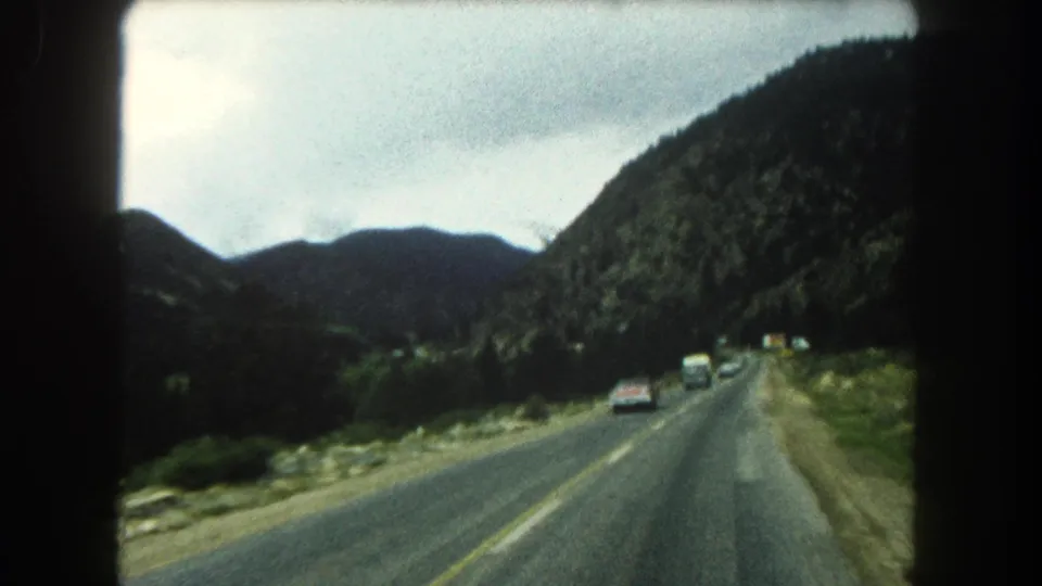 A moving-car view of a Summit County highway in 1962, with several vehicles ahead and dark green mountain slopes beyond.