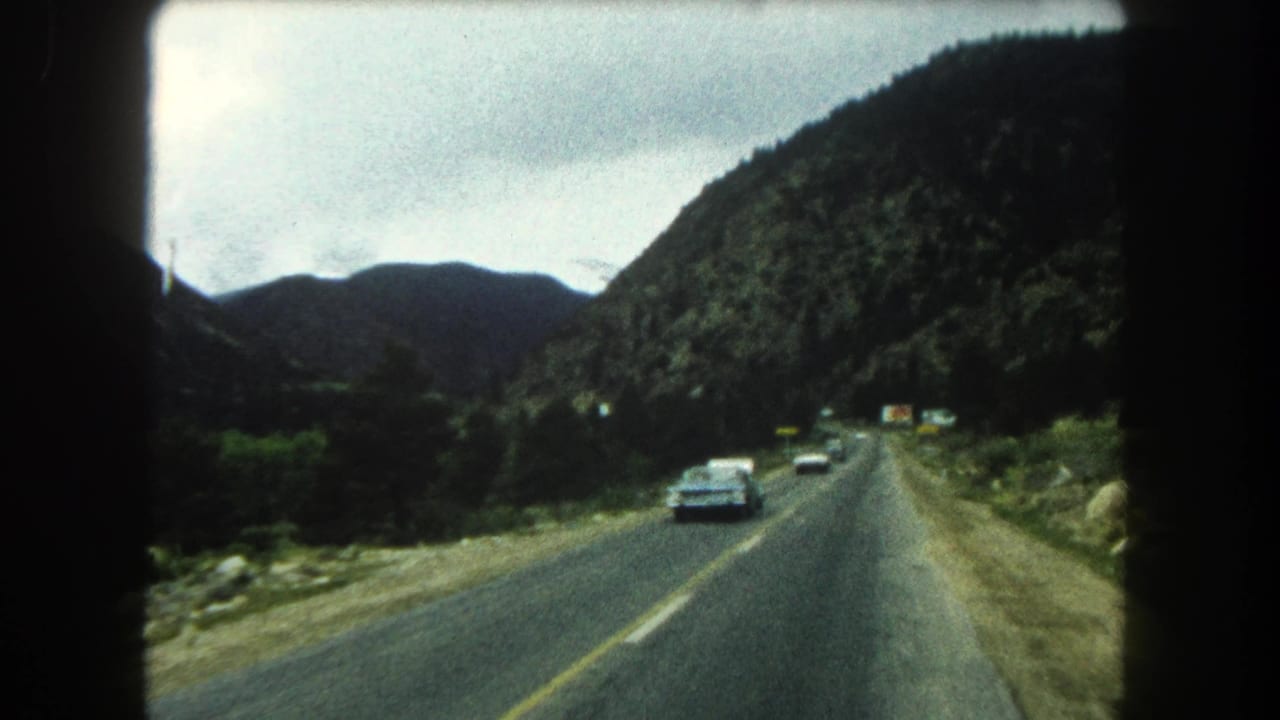 Two-lane mountain highway with oncoming cars, forested slopes, and an overcast sky.