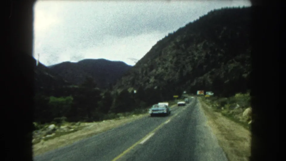 1962 Summit County, Colorado home-movie view from a moving vehicle on a two-lane mountain highway with oncoming cars and wooded slopes.