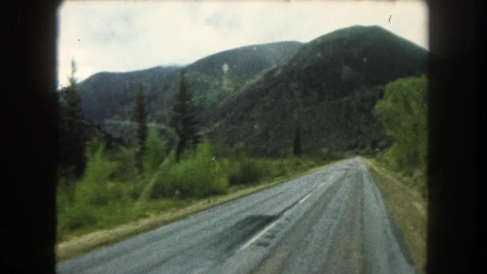 Moving 8mm view of a Summit County, Colorado mountain road in 1962, with pavement, roadside trees, brush, and wooded hills ahead.
