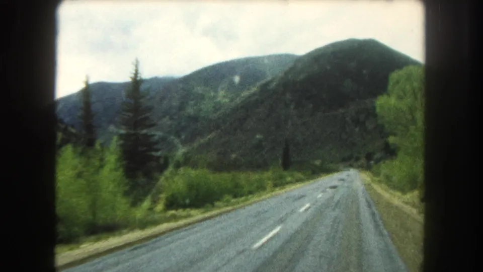 1962 Colorado home-movie frame of a two-lane mountain road bordered by trees and brush, with forested slopes under a pale cloudy sky.