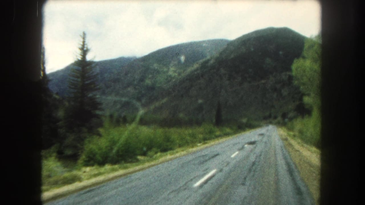1962 Summit County, Colorado home-movie frame looking through a car windshield at a paved road, roadside trees, and forested mountain slopes.