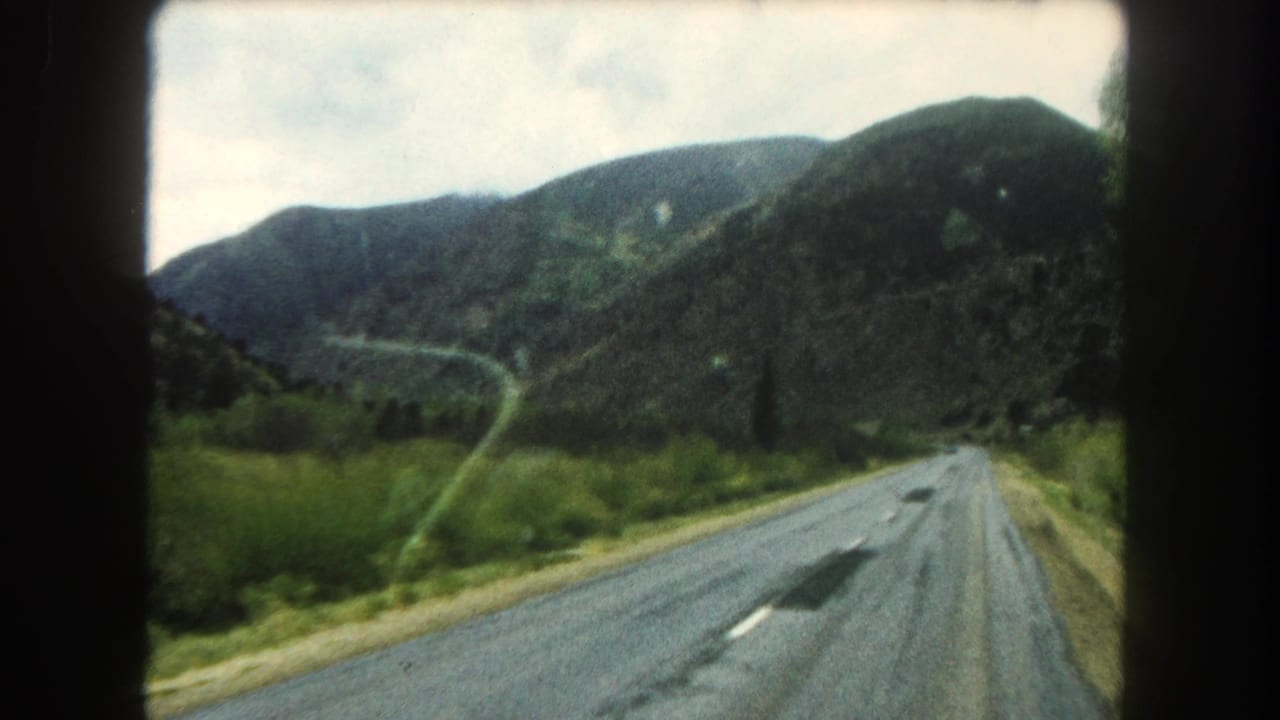 Winding road through green mountain terrain, filmed from a car with dark frame edges visible.