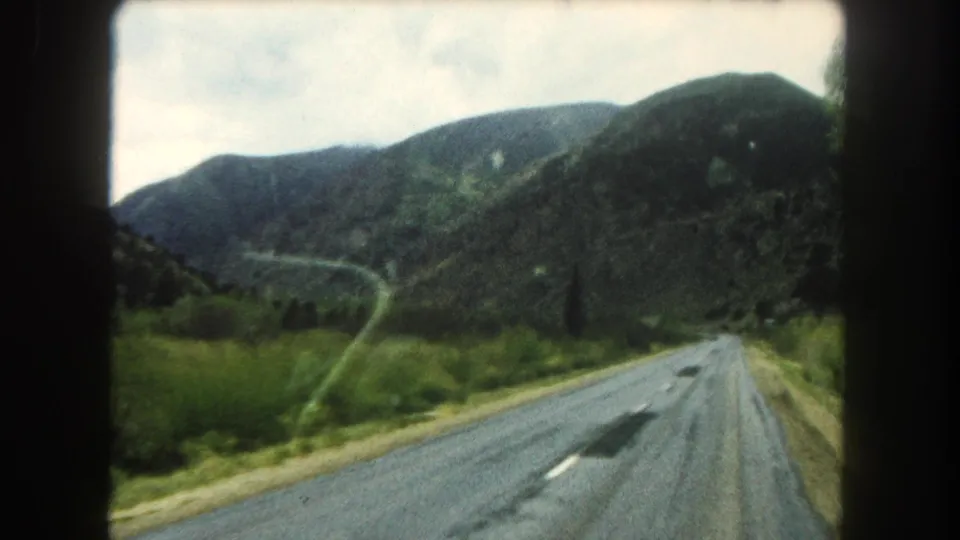 In-car 8mm view of a Summit County mountain road in 1962, with a distant curve, forested hills, and soft motion blur.