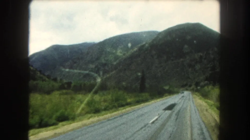 1962 Colorado road-travel frame showing an asphalt road, a dark roadside tree, and layered green mountain slopes from a moving car.