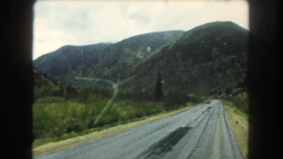 Summit County, Colorado home-movie frame from 1962 showing a curving mountain road, green hills, and cloudy sky from inside a moving car.