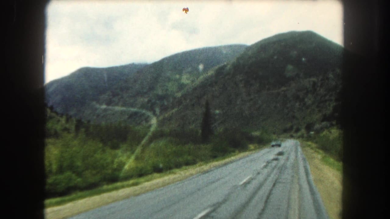 1962 8mm view of a two-lane road through Summit County mountain terrain, with wet pavement, green roadside vegetation, and film-gate borders.