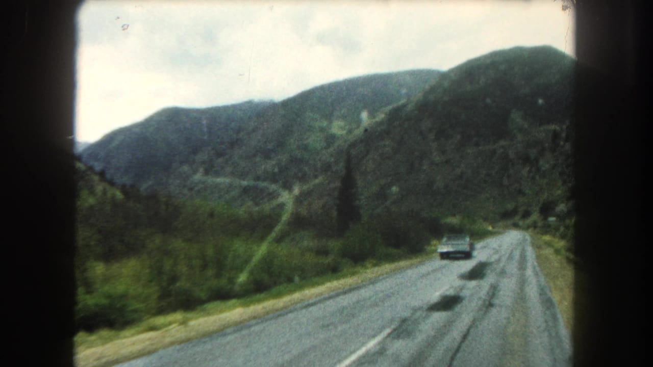 A mid-century car appears on a wet two-lane road in Summit County, Colorado, with green hills, mountains, and dark 8mm frame edges visible.