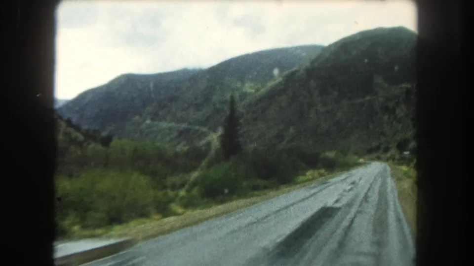 Home-movie frame of a wet two-lane road curving through Summit County hills, with dense roadside plants, mountain slopes, and soft 8mm color.