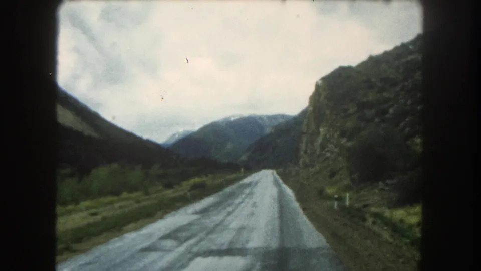 A Summit County two-lane road stretches between rocky mountain sides and distant hills, captured with soft 1962 8mm home-movie texture.