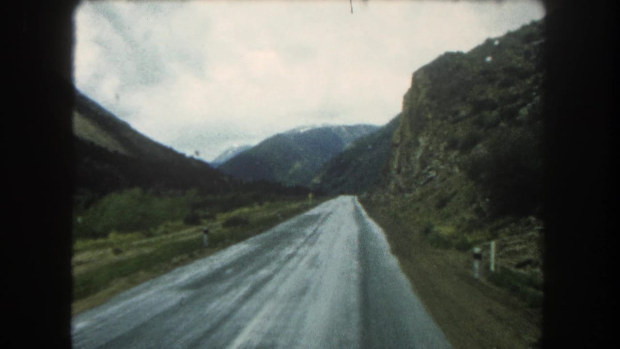 1962 Summit County travel view from a moving vehicle on a wet road beside rocky mountain slopes and distant peaks.