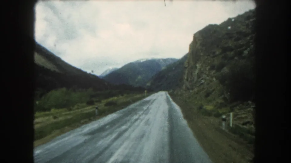 1962 Summit County travel view from a moving vehicle on a wet road beside rocky mountain slopes and distant peaks.