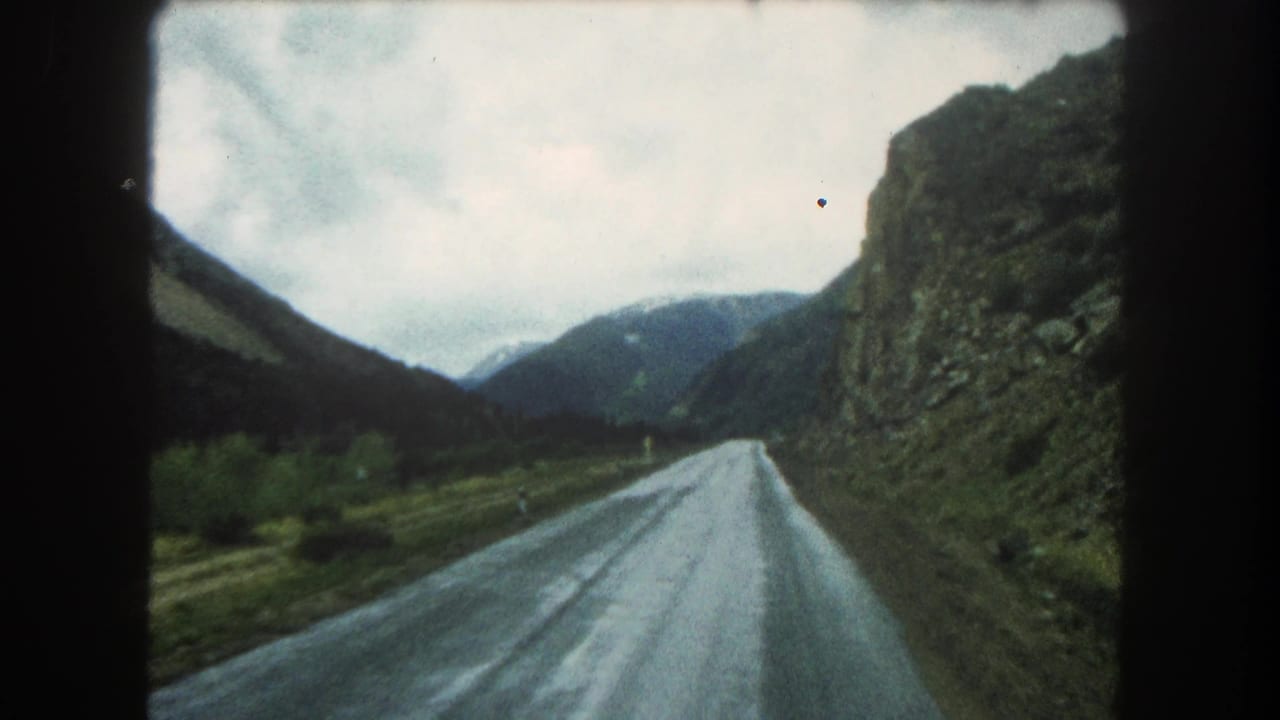 Moving 8mm view in Summit County, Colorado, showing a wet mountain road bordered by a rock cut and dark slopes.