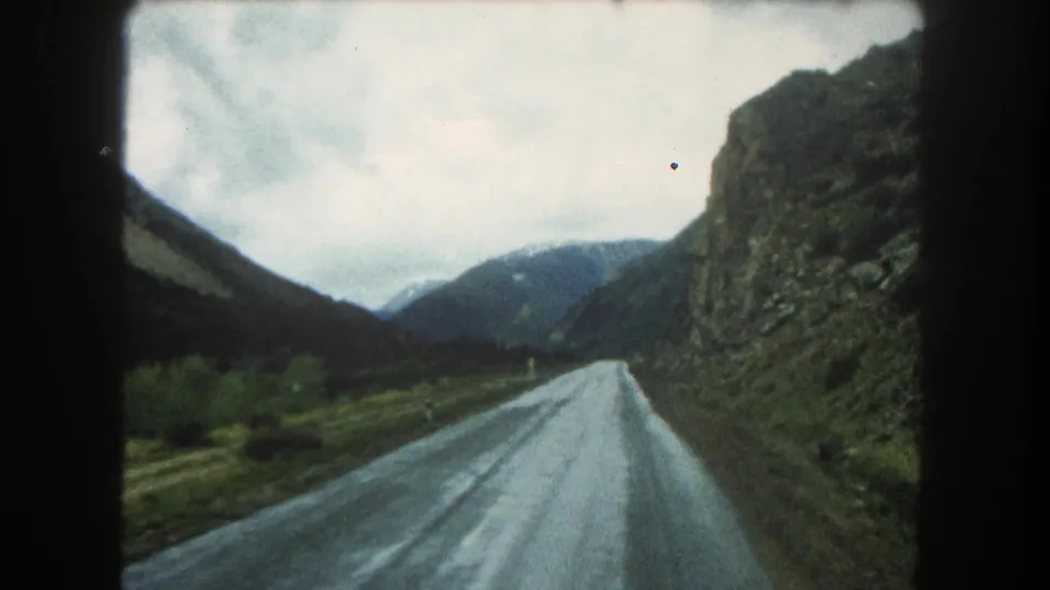 Moving 8mm view in Summit County, Colorado, showing a wet mountain road bordered by a rock cut and dark slopes.