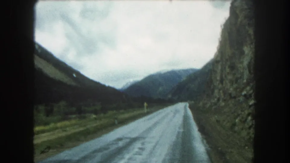 Final 1962 Colorado travel frame showing the wet road narrowing toward distant mountains beside a rugged rock face.