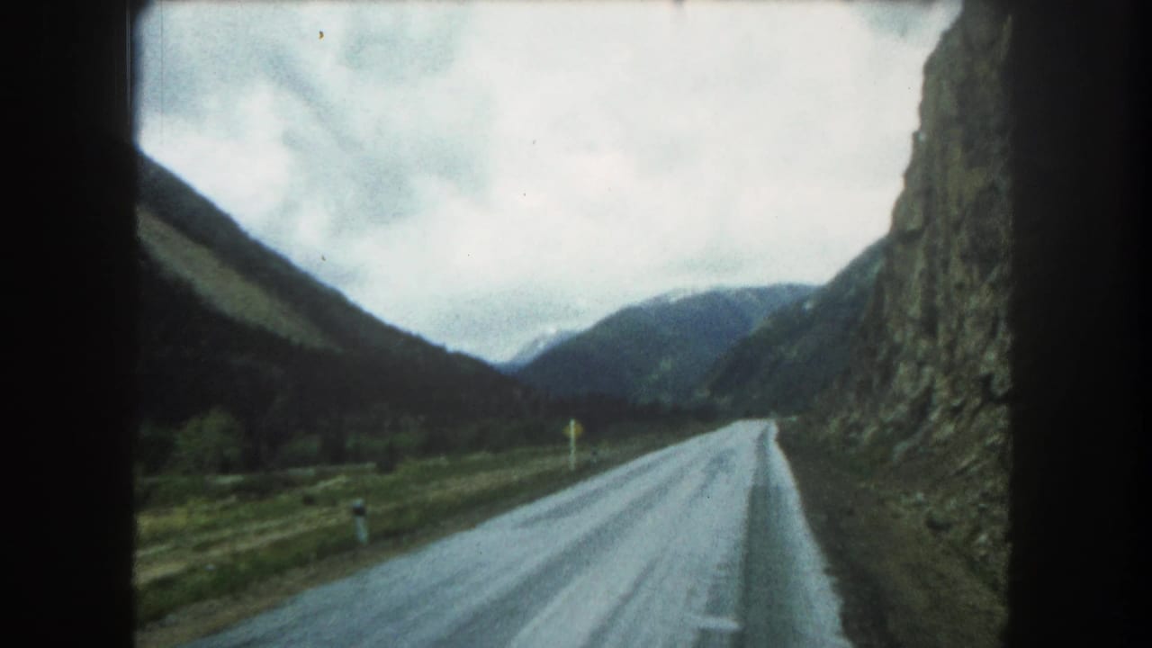 1962 8mm home-movie frame looking down a wet Summit County mountain road with steep slopes and an overcast sky.