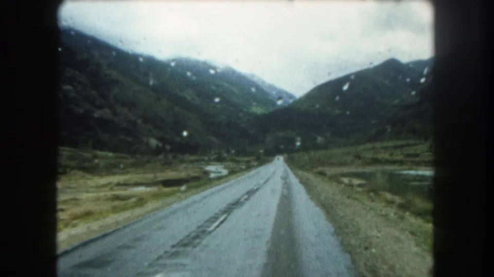Rain-speckled 1962 8mm frame showing a Summit County road descending between dark mountain slopes and wet ground.