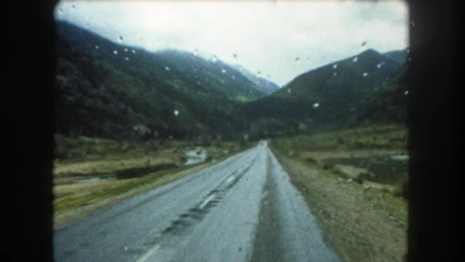 1962 Summit County driving POV with raindrops on the lens, a wet road, and green mountain slopes ahead.