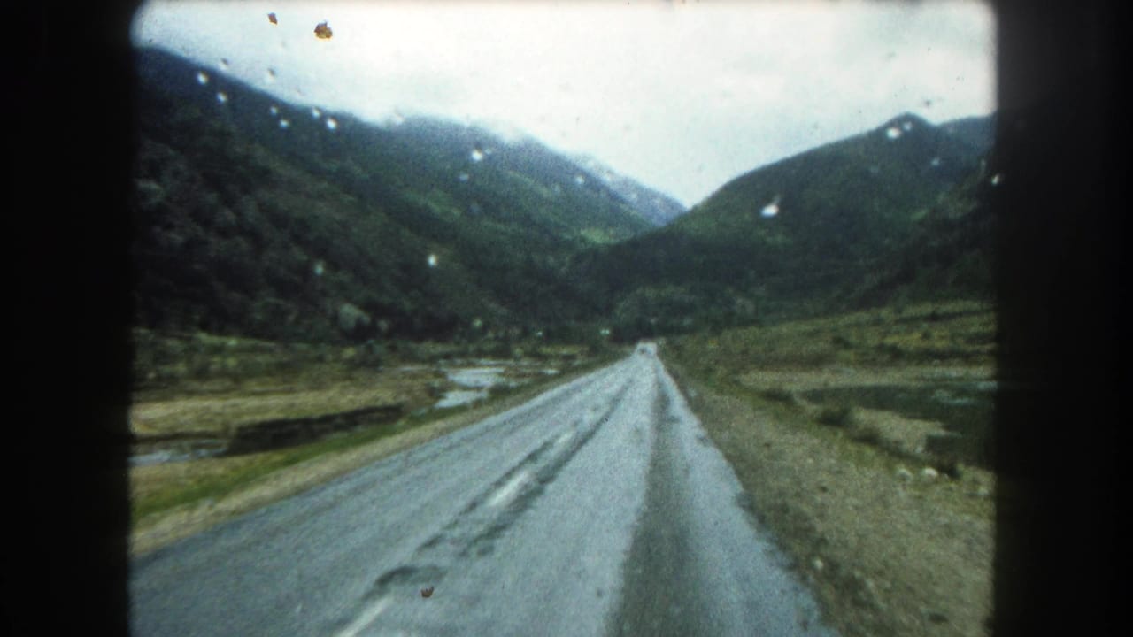 1962 Summit County, Colorado home-movie frame showing a wet road receding through a green mountain valley under overcast light.