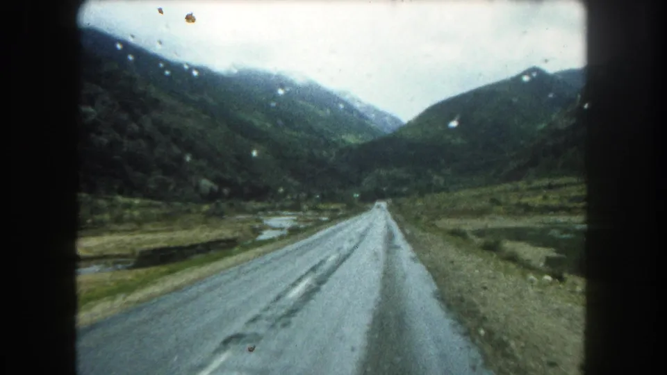 1962 Summit County, Colorado home-movie frame showing a wet road receding through a green mountain valley under overcast light.