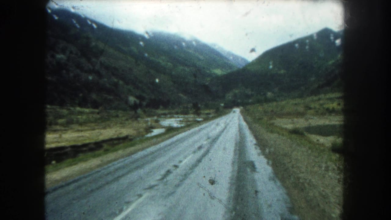 A wet paved road stretches ahead through a Colorado mountain valley, with raindrops and film gate borders visible in the 8mm frame.