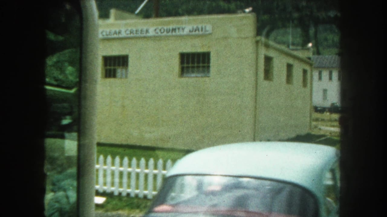 1962 8mm frame showing the Clear Creek County Jail exterior, barred windows, white picket fence, and a partial car at the roadside.