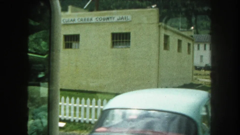 1962 8mm frame showing the Clear Creek County Jail exterior, barred windows, white picket fence, and a partial car at the roadside.