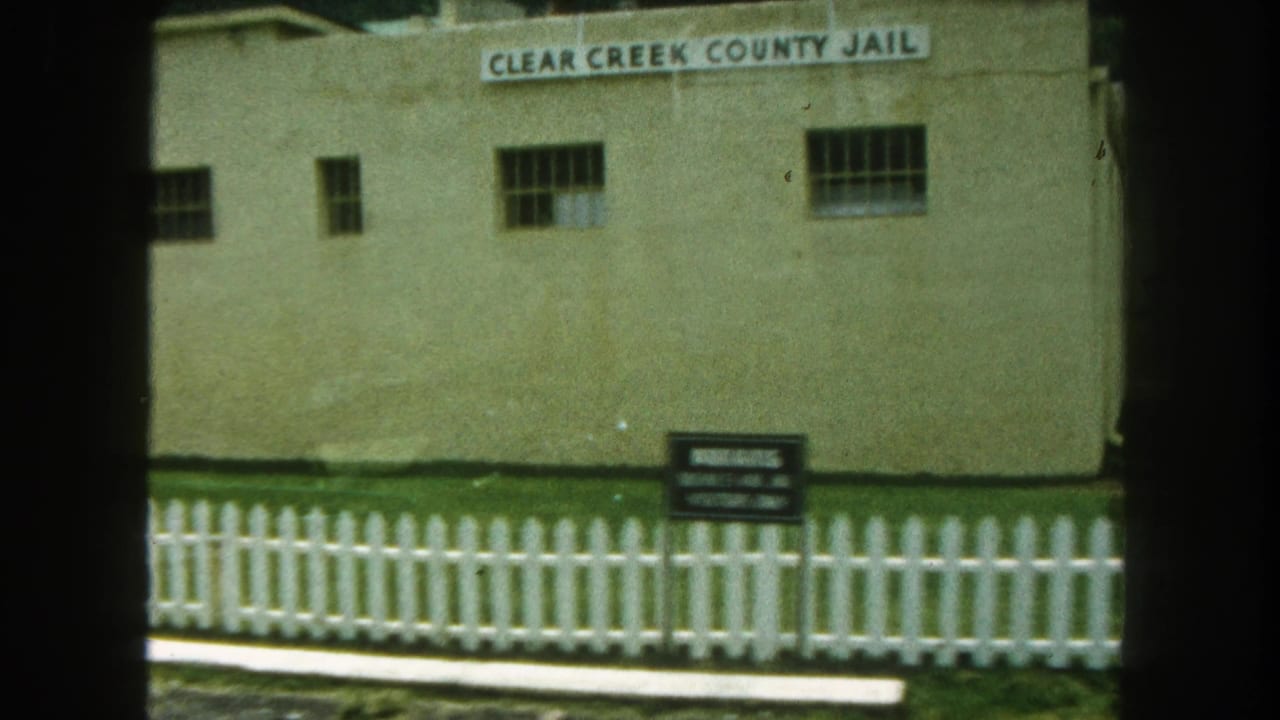 1962 home-movie frame of the Clear Creek County Jail building, with name sign, barred windows, lawn, and white fence visible.