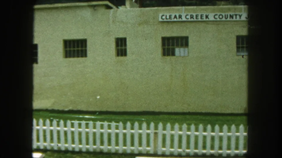 1962 8mm frame cropping across the Clear Creek County Jail facade, showing barred windows, signage, and the picket fence below.
