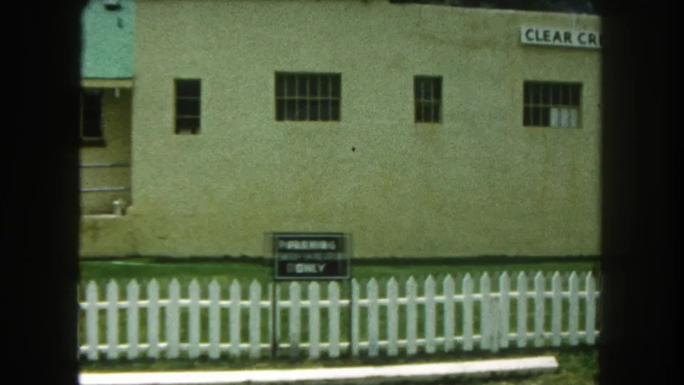 1962 Summit County, Colorado home-movie frame showing a beige institutional-looking building, white picket fence, and parking sign.