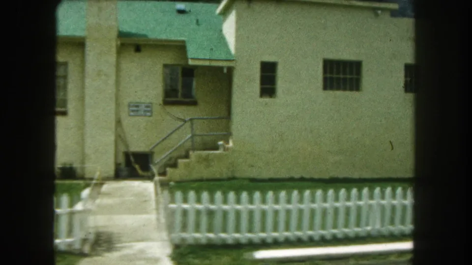 1962 Summit County, Colorado frame with a beige building, green roof, exterior stairway, walkway, lawn, and white fence.