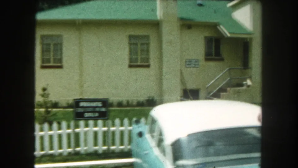 1962 Summit County, Colorado frame showing a period car passing the fenced beige building and exterior stairway.