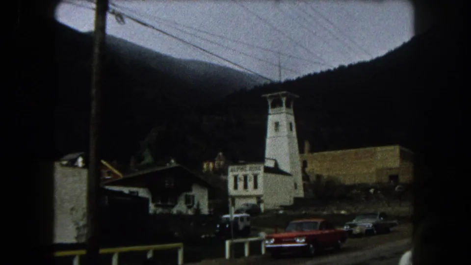 1962 Summit County, Colorado home-movie frame showing a mountain street, white tower-like building, vintage cars, utility lines, and dark slopes.
