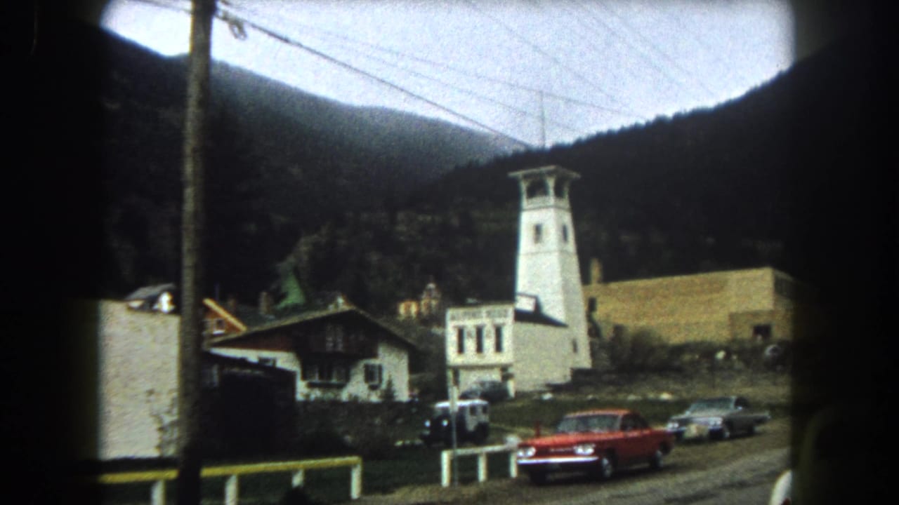 Mountain town roadside with a white tower, Alpine Hotel sign, red car, utility pole, and forested slopes.