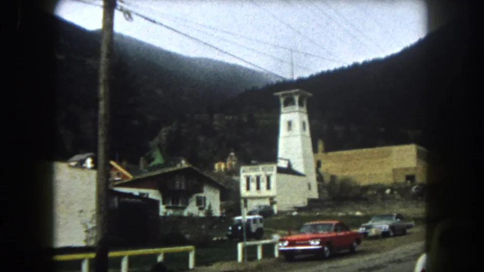 1962 Summit County, Colorado roadside view with a white tower, Alpine Hotel sign, red sedan, parked cars, power lines, and mountains.
