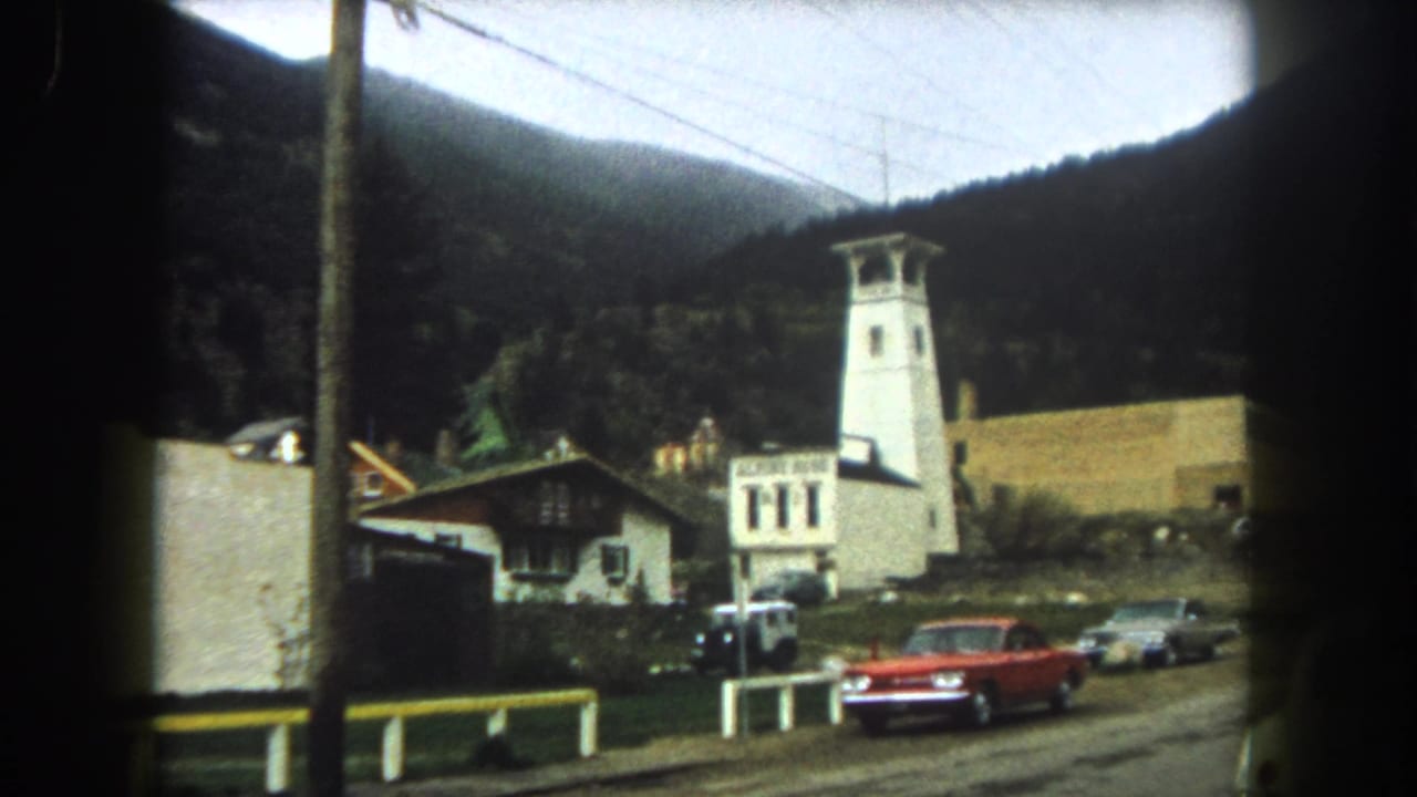 White tower and hotel sign near a roadside, with a red sedan, parked cars, utility lines, and mountains.