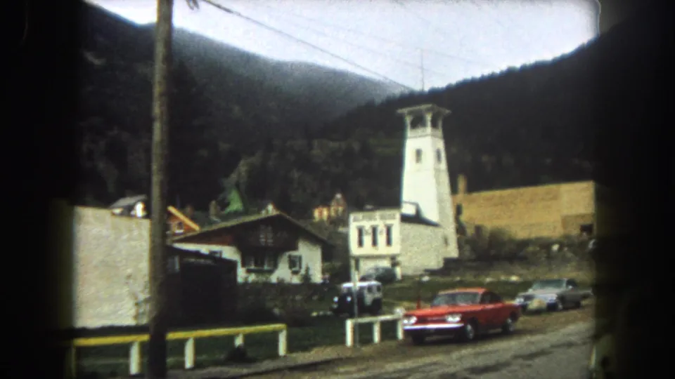 1962 home-movie frame of a Summit County roadside hotel area, with tower, Alpine Hotel sign, red sedan, cars, and forested mountains.
