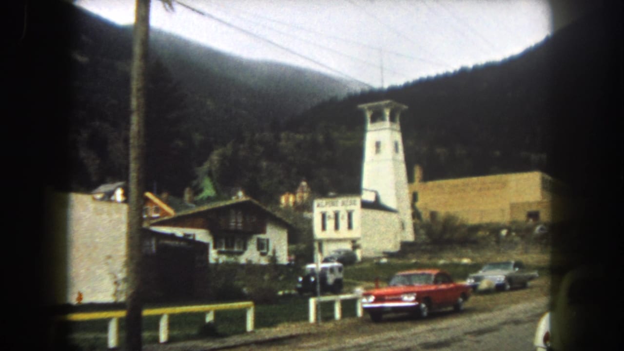 1962 Summit County street view with a red car, white tower-like building, roadside buildings, power lines, and forested mountains.