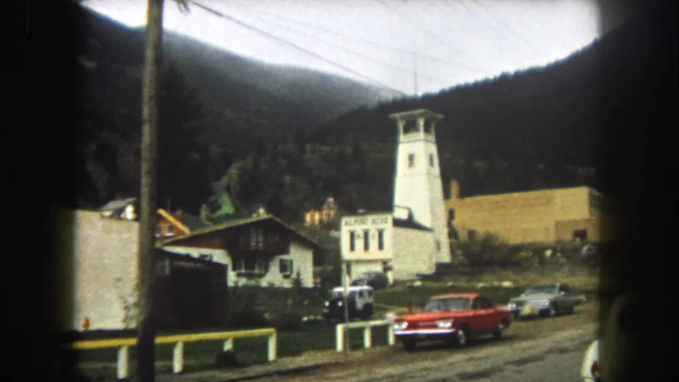 1962 Summit County street view with a red car, white tower-like building, roadside buildings, power lines, and forested mountains.
