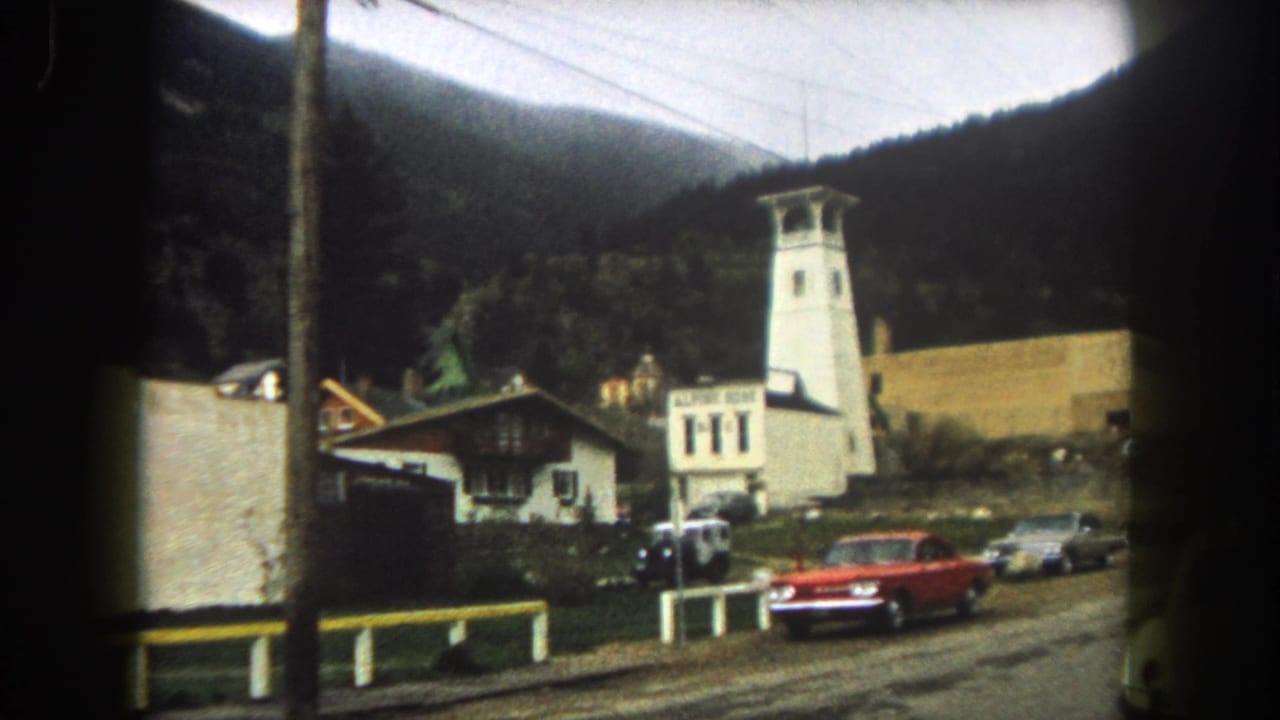 Red car on a mountain street near a white tower building, roadside structures, and overhead wires.