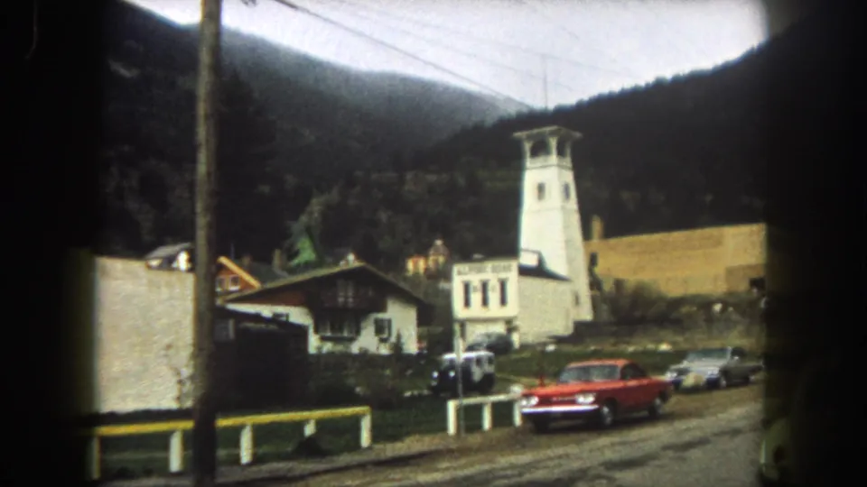 1962 Summit County street scene with a red car, white tower-like building, small roadside buildings, overhead wires, and mountain slopes.