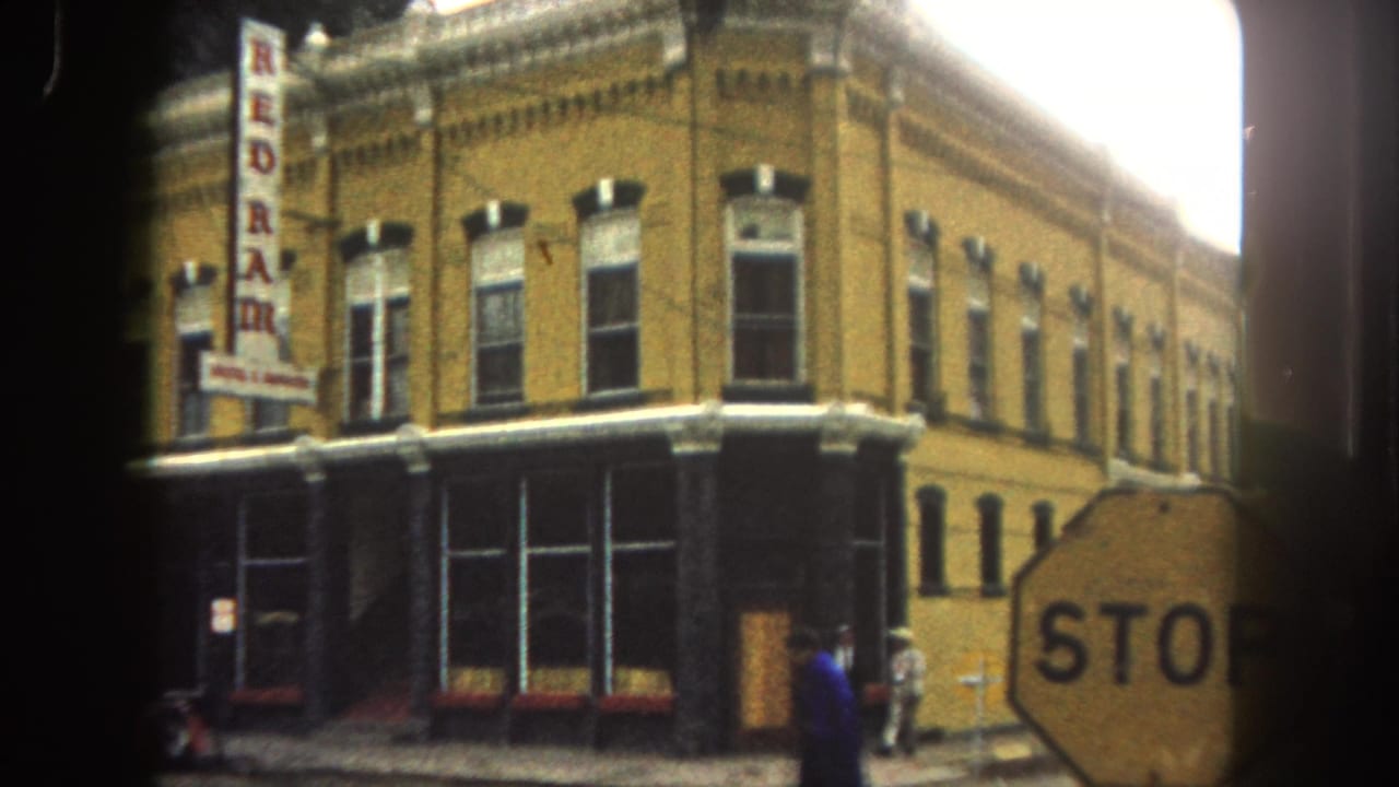 1962 Summit County street corner with a yellow brick building, vertical REDRAM sign, stop sign, and pedestrians passing the storefront.