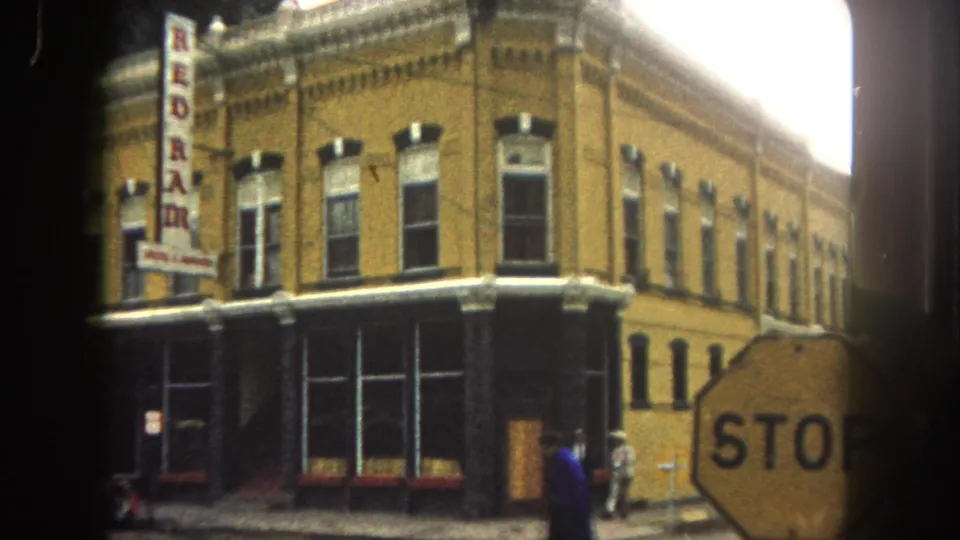 1962 Summit County street corner with a yellow brick building, vertical REDRAM sign, stop sign, and pedestrians passing the storefront.